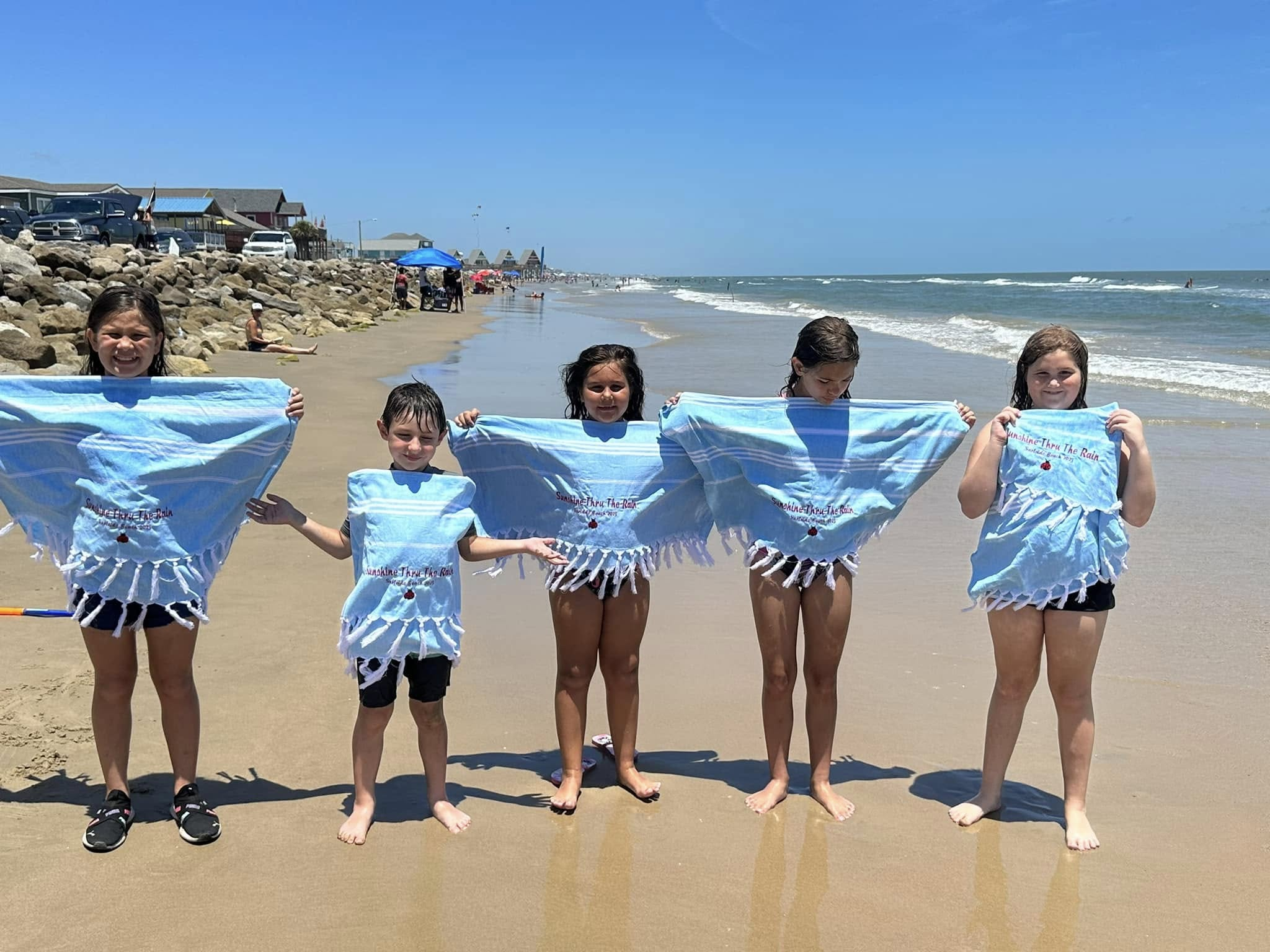 Children stand together on the beach holding blue towels