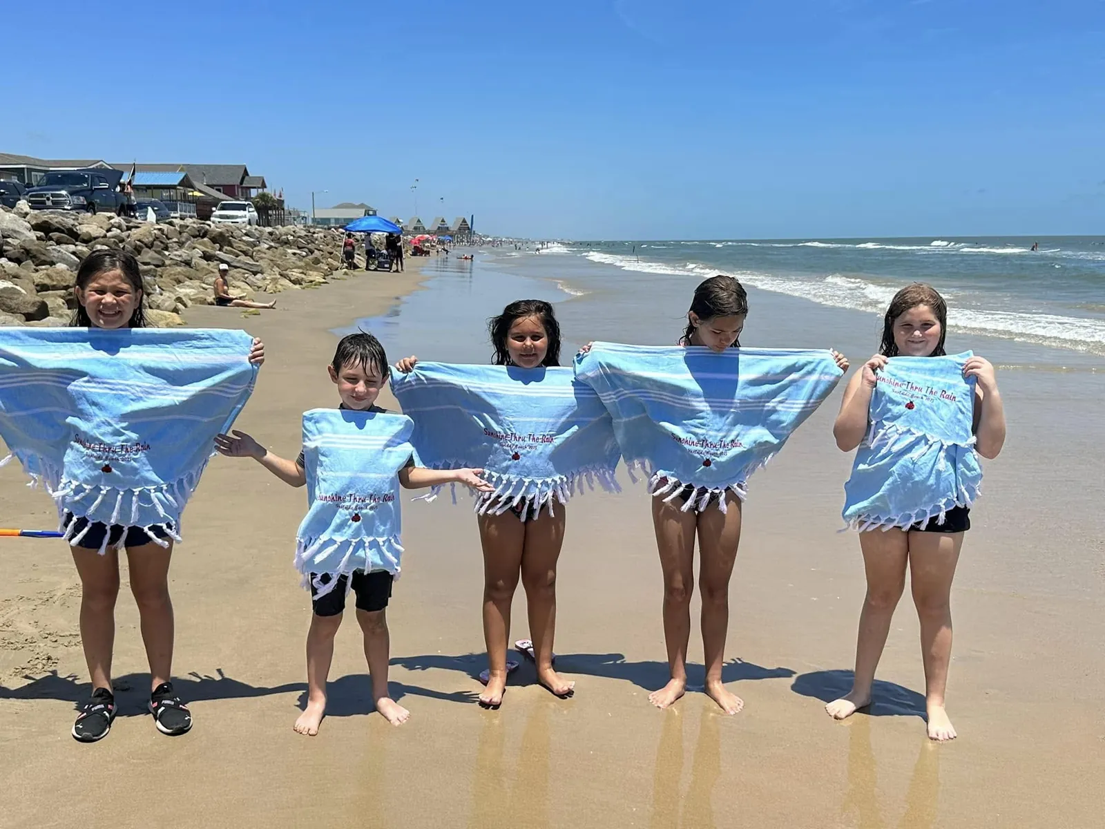 Children stand together on the beach holding blue towels