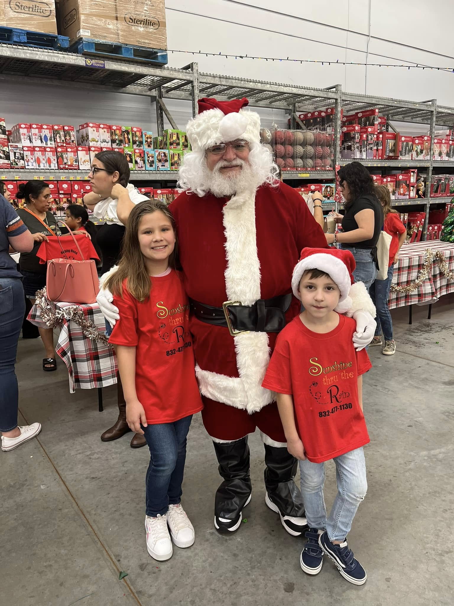 Two children in Sunshine Thru the Rain shirts smile beside Santa during a holiday event
